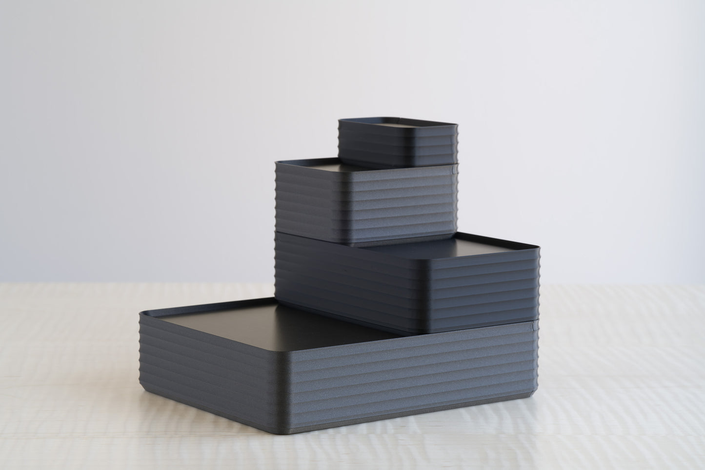 Four black and metallic iron gray ribbed boxes stacked in a tiered arrangement on a maple wood table, modern minimalist storage design.