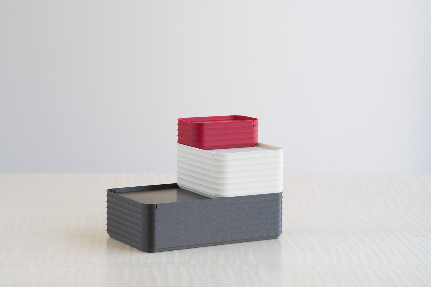 Trio of stacked ribbed boxes in metallic iron gray, bone white, and crimson red, displayed on a maple table for bold contrasting design appeal.
