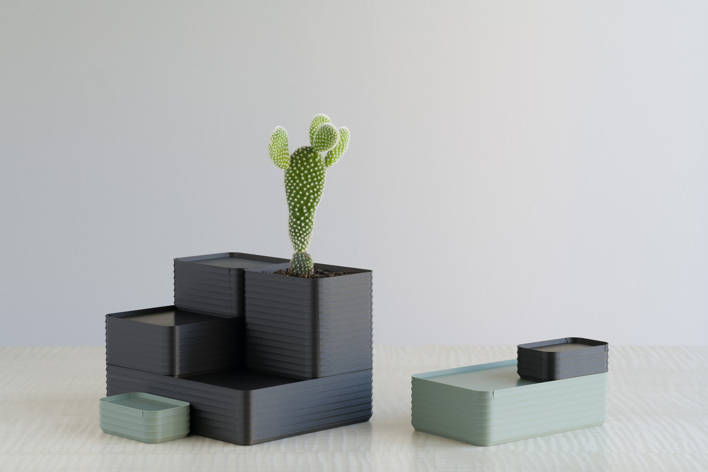 Two sets of Z Co Rib Series boxes on a maple table, featuring metallic iron gray and muted green tones. One stack includes a cactus planted in a planter box, showing the multifunctional use of the ribbed containers as planters or storage solutions.