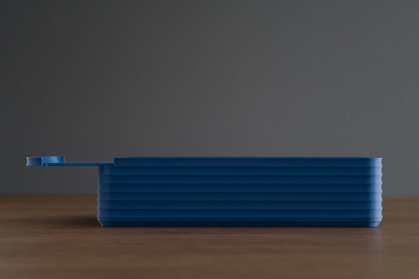 Side profile of the Z Co Rib Series Double-Wide Box in matte dark blue, placed on a walnut surface. The sliding lid is partially open, showing the ribbed texture and elongated rectangular design.