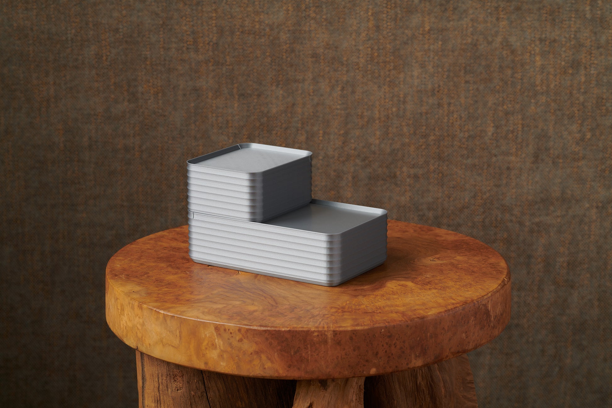 Original size silver Rib Series Boxes styled arrangement photo in a stepped stack on a round wood stool with warm brown background, highlighting minimalist design.