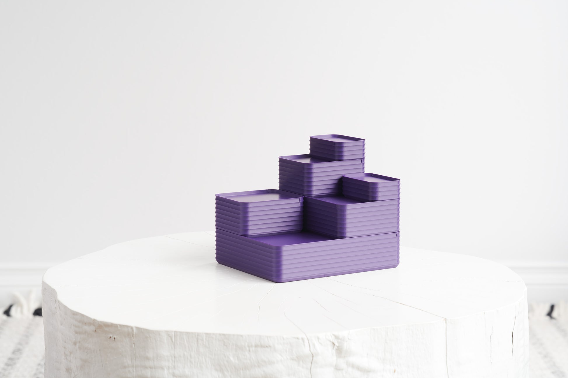 A stepped arrangement of six Z Co Rib Series boxes in dark violet, neatly stacked on a white round stool against a minimalist white background.