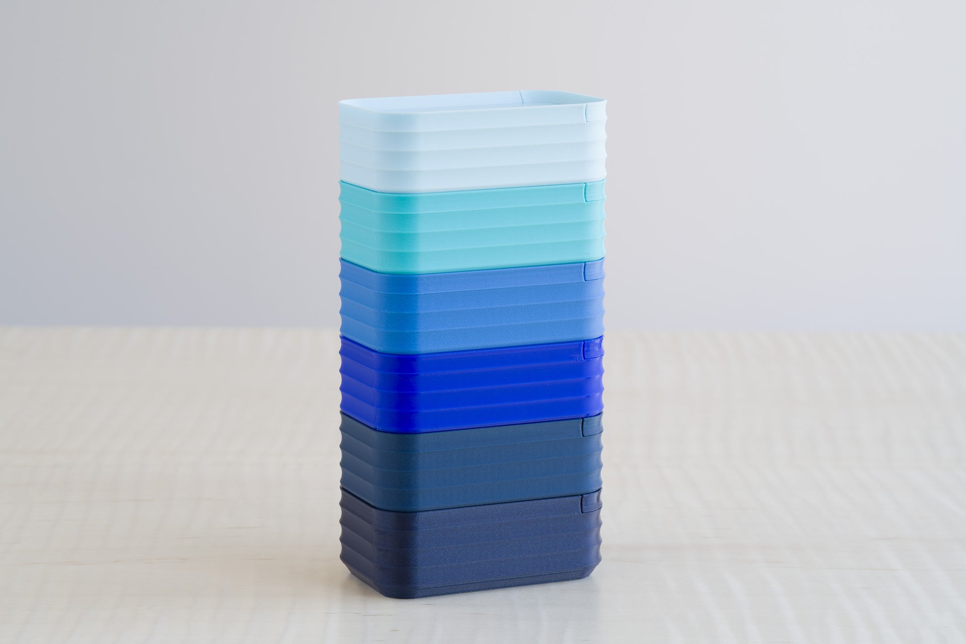 Stacked Rib Series Mini Boxes in gradient blue tones, including matte ice blue, turquoise, matte blue, cobalt blue, matte dark blue, and metallic dark blue, arranged vertically on a light wood table.