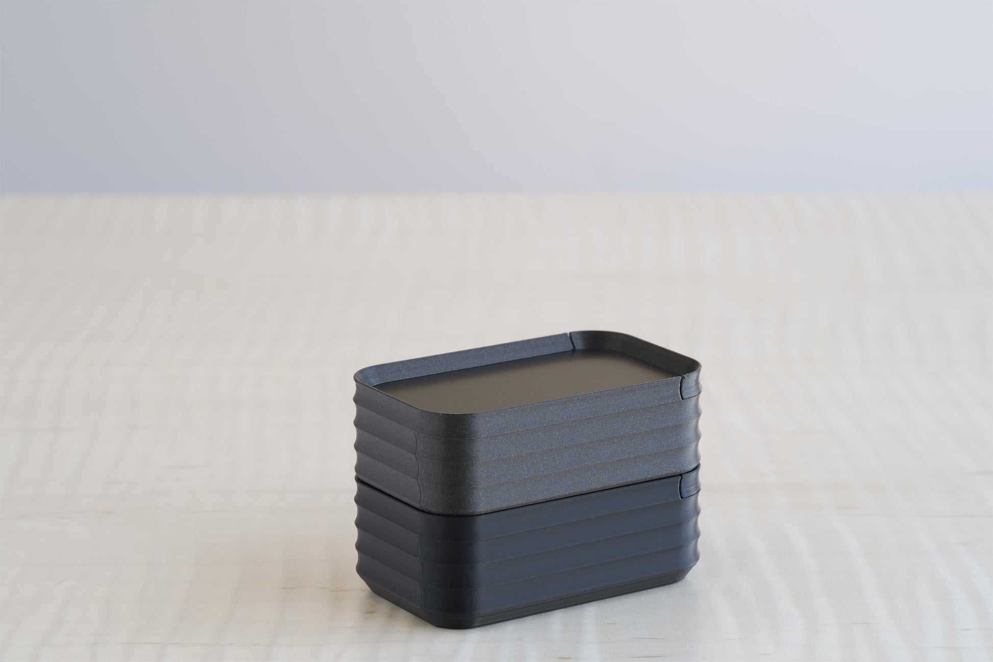 Pair of Rib Series Mini Boxes in metallic iron black and black, stacked neatly on a light wood table to showcase dark neutral finishes.